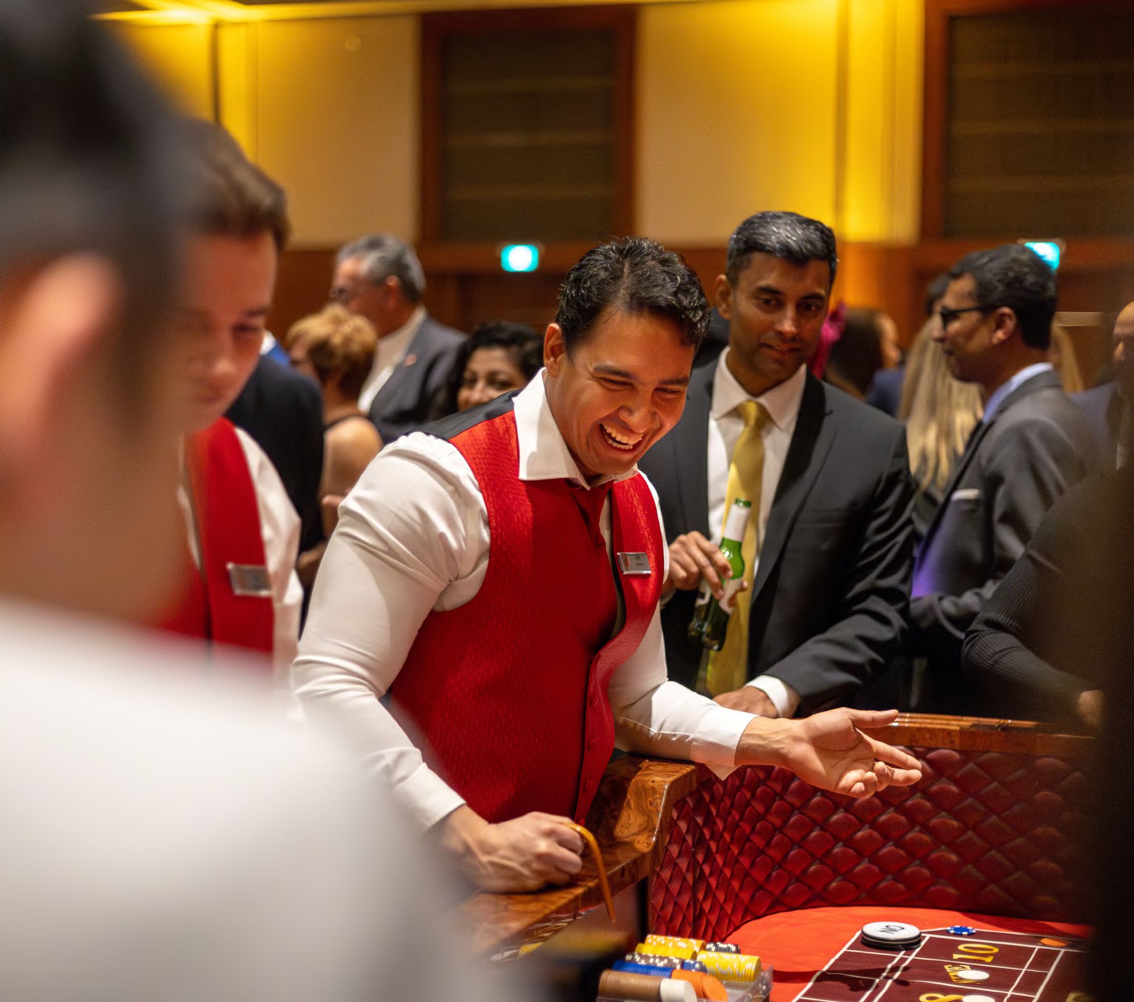 A dealer in a red vest laughing with guests around a casino table, photographed by Dario Hudon